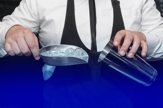 The hands of a professional bartender add ice in a scaple to a tool for preparing and mixing alcoholic cocktails, a metal shaker.