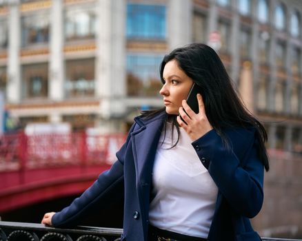 Woman with long hair talking on phone. Autumn or winter, girl in outdoor. Beautiful intelligent brunette in street of Saint-Petersburg in a city center near bridge, copy space
