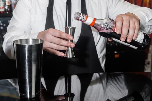 The hands of a professional bartender pour red syrup into a measuring glass of jigger, next to a metal tool for preparing and stirring alcoholic cocktails of shaker drinks.