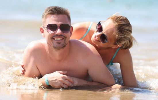 Portrait of smiling couple laying on coastline, posing for memorable picture from vacation. Middle aged man and pretty woman in sunglasses. Love concept