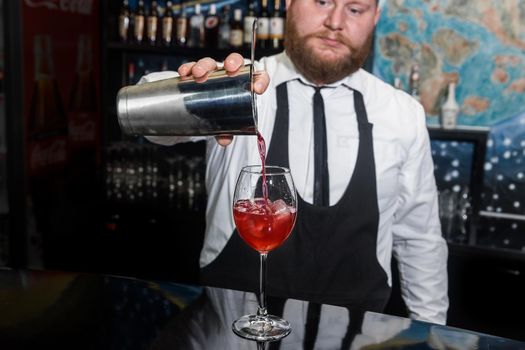 Professional bartender with a beard pours liquid into a glass of ice cubes from a tool for mixing and preparing alcoholic beverages cocktails metal shaker.