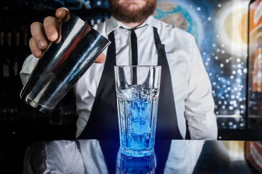 The process of preparing a cocktail. The hand of a professional bartender holds a metal shaker liquid mixing tool next to a glass with ice and blue syrup.