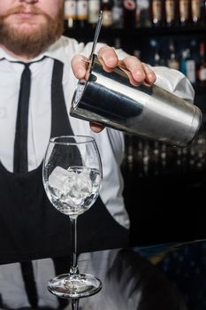 Professional bartender with a beard pours liquid into a glass of ice cubes from a tool for mixing and preparing alcoholic beverages cocktails metal shaker.