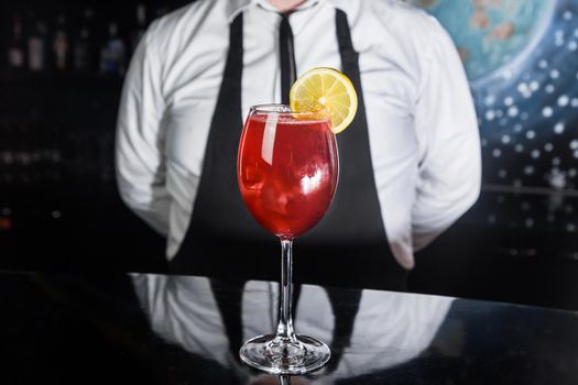 Red alcoholic beverage cocktail in glass decorated with a slice of lemon on the bar counter against the background of the bartender.