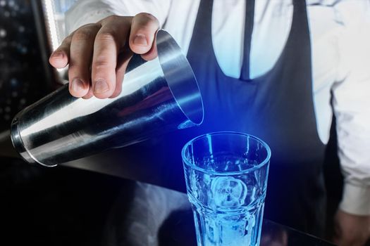 The hand of a professional bartender holds a tool for mixing and making alcoholic cocktails, a metal shaker and pours blue syrup into an ice glass.