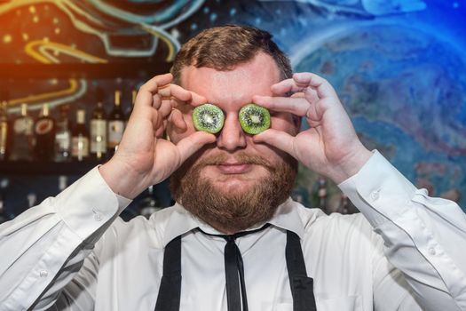 Adult funny bearded positive man professional bartender holds halves of sliced kiwi in his hands at the face.