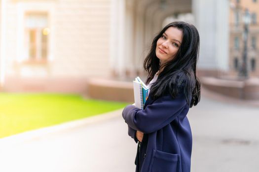 Beautiful smiling girl brunette student holding notebooks and textbooks, stands at University on street of St. Petersburg. A charming woman with long dark hair is studying at a course, copy space