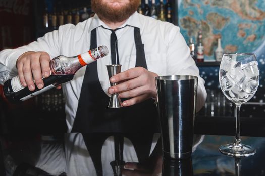 The hands of a professional bartender pour red syrup into a measuring glass of jigger, next to a metal tool for preparing and stirring alcoholic cocktails of shaker drinks and glasses with ice cubes.