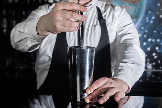 The hands of a professional bartender interfere with a bar spoon the contents of a metal shaker tool for preparing and stirring alcoholic cocktails.