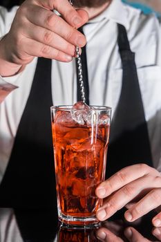 The hand of a professional bartender stirs the red syrup in an alcoholic cocktail with a bar spoon on the bar counter. The process of preparing an alcoholic beverage.
