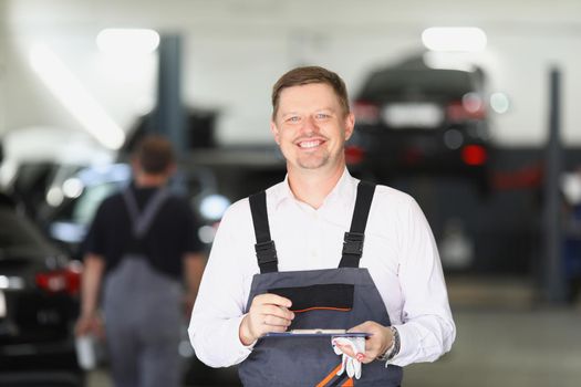 Portrait of smiling maintenance center worker in uniform, man fill in papers on car checkup. Mechanic male propose high service. Pit stop, garage concept