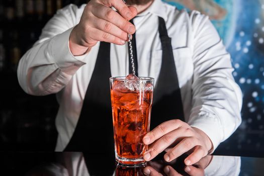 The hand of a professional bartender stirs the red syrup in an alcoholic cocktail with a bar spoon on the bar counter. The process of preparing an alcoholic beverage.