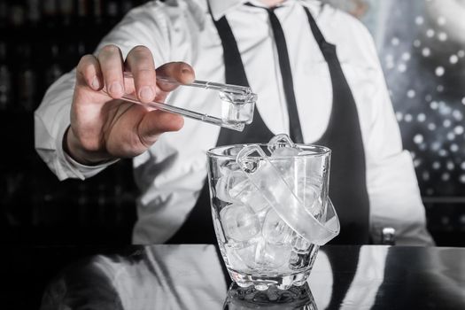 The hand of a professional bartender holds plastic ice cube tongs next to a cold ice dish.