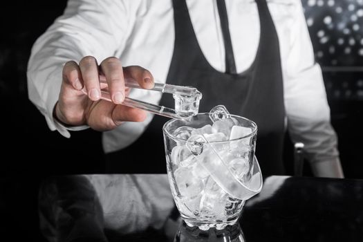 The hand of a professional bartender holds plastic ice cube tongs next to a cold ice dish.