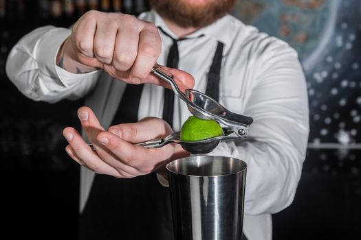 Hands of a professional bartender squeeze lime juice with an iron tool with a juicer into a metal shaker, close-up.