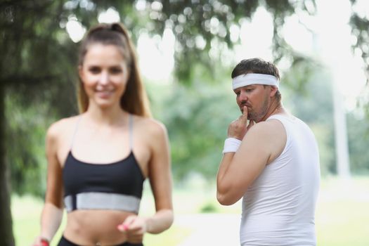 Portrait of middle aged man check on young beautiful fit woman running in park. Active woman jogging in front, feeling energetic. Sport, health concept