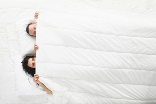 Top view of woman and man peeking on each other under white blanket, awake couple in bed. Good morning, play, fun, family, chilling, sweet home concept