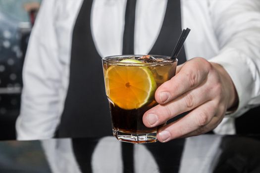 The hand of a professional bartender serves an alcoholic beverage cocktail whiskey with cola and a piece of lime on the bar counter, close-up.