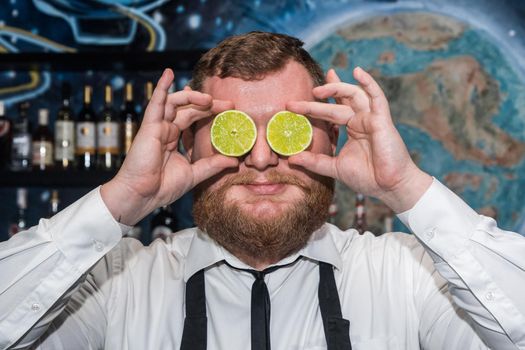 Adult funny bearded positive man professional bartender holds halves of sliced lime in his hands at the face.