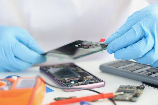 Close-up of handyman repairing broken smartphone, put cover on top, worker wear gloves. Workplace full with equipment. Repair service, fix, handy concept