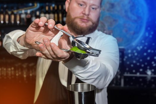 Hands of a professional bartender squeeze lime juice with an iron tool with a juicer into a metal shaker, close-up.