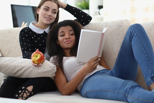 Portrait of best friends reading book chilling on sofa, apple for snack, relaxing atmosphere. Expand knowledge in literature. Leisure, friendship concept