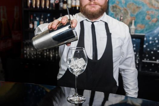 Professional bartender with a beard pours liquid into a glass of ice cubes from a tool for mixing and preparing alcoholic beverages cocktails metal shaker.