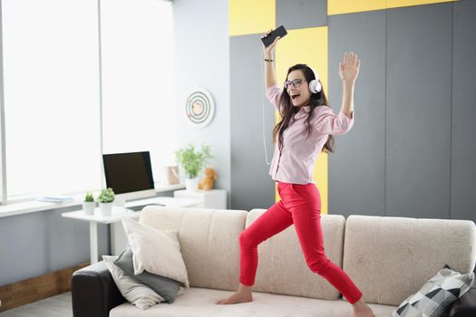 Portrait of happy woman dancing on couch, listen to music in headset and sing. Happy mood, good news, celebrate, party. Positivity, fun, go nuts concept