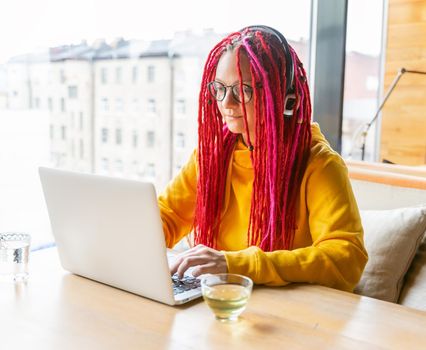 Woman in headphones is looking at a laptop and plays a computer game over the network. Girl with long pink hair. designer, creative specialist, freelancer, journalist. Digital Nomad Concept