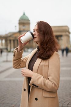 Beautiful serious stylish fashionable smart girl drinks coffee, goes walking down street of St. Petersburg in city center. Charming thoughtful woman with long dark hair, vertical