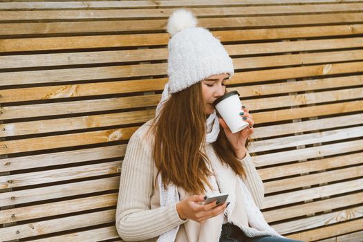 Beautiful young girl drinking coffee, tea from a plastic mug in autumn, winter and talking on a mobile phone. Woman with long hair sitting on a bench in autumn or winter, basking in a hot drink, copy space