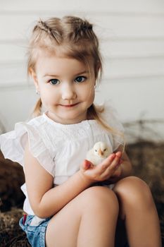Selective focus of lovely girl with big beautiful eyes looking at camera while keeping little chick in hands. Cute child smiling and posing while resting in village. Concept of childhood and animals.