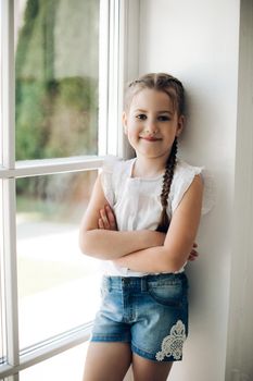 Portrait of little cute kid leaning on wall and looking at camera. Small pretty girl standing near big window and smiling. Adorable child with braid in casual clothes holding arms crossed on chest.