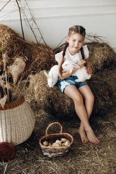 Portrait of cute little girl with two braids sitting on stack of hay with pet white rabbits. She is looking at camera hugging one rabbit.