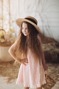 Portrait of little charming kid in peachy elegant dress and hat shyly posing at camera. Small cute girl standing among decoration and bunches of hay. Sweet lovely child with long hair holding hand up.