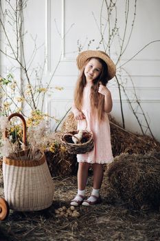 Portrait of little charming kid in peachy elegant dress and hat shyly posing at camera. Small cute girl standing among decoration and bunches of hay. Sweet lovely child with long hair holding hand up.