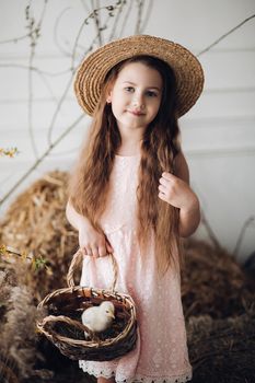 Front view of beautiful girl wearing pink dress and hay hat keeping basket with little chick. Cute female child looking aside and smiling while walking in garden in village. Concept of childhood.