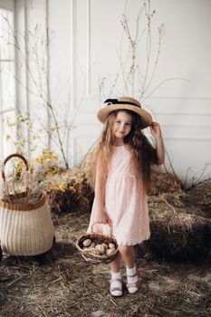 Portrait of little charming kid in peachy elegant dress and hat shyly posing at camera. Small cute girl standing among decoration and bunches of hay. Sweet lovely child with long hair holding hand up.