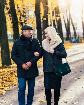 Happy blonde mature woman and handsome middle-aged brunette man walk in park, looking at each other. A loving couple of 45-50 years old walks in the autumn park in warm clothes