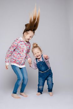 Front view of two pretty sisters keeping hands, looking at camera and smiling on isolated background in studio. Happy kids wearing stylish outfits posing together. Concept of friendship and fashion.