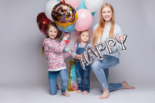 Front view of happy family looking at camera and posing while celebrating birthday party. Young mother keeping little daughter while girl standing near and keeping colorful balloons. Concept of fun.