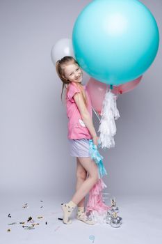 Studio portrait of lovely little girl with red or fair hair and freckles in pink t-shirt and shorts posing with toothy smile with pink, blue and white air balloons against grey background.