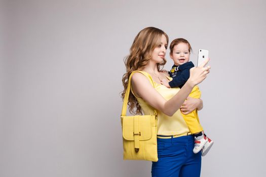 Side view of pretty mother walking with little kid and taking selfie. Young woman keeping child on hands and posing on isolated background in studio. Concept of childhood and family outfit.