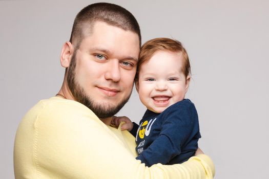 Portrait of happy father in yellow sweater posing with his lovely smiling child. Athletic and young men with beard holding red haired little boy. Handsome brunette dad hugging sweet and cute son.