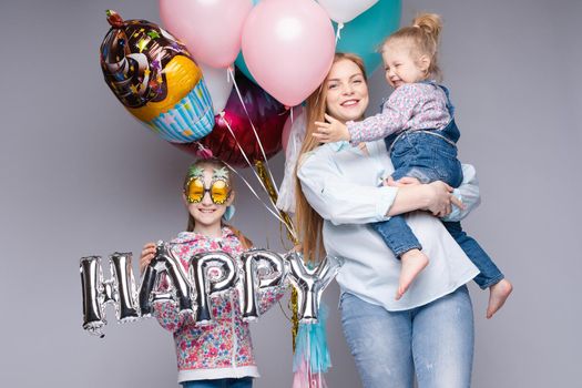 Front view of happy family looking at camera and posing while celebrating birthday party. Young mother keeping little daughter while girl standing near and keeping colorful balloons. Concept of fun.