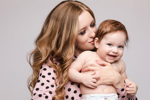 Portrait of lovely mother kissing little child on isolated background in studio. Pretty woman keeping cheerful daughter on hands and hugging her. Concept of family love and childhood.