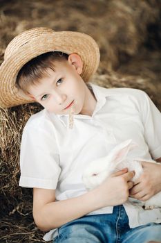 Front view of boy wearing white shirt and hay hat keeping cute white bunny and smiling. Young male hugging rabbit and posing while resting in village. Concept of childhood, animals and love.