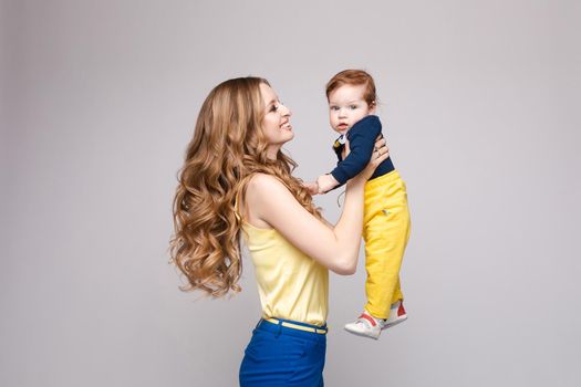 Young and fit woman in casual clothes holding little kid. Beautiful mother in yellow blouse and blue pants posing from back with her lovely son. Blonde mom on heels looking and happy smiling child.