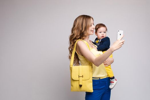 View from side of beautiful mother with little child keeping phone and doing selfie on grey isolated background. Happy family in yellow outfit smiling and posing in studio. Concept of technology.