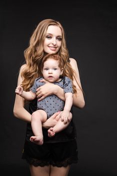 Front view of positive little baby laughing with mother in studio. Beautiful woman in black pajamas keeping cute daughter on hands. looking at camera and posing on black isolated background.